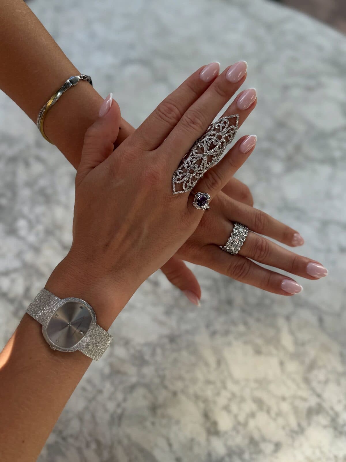 Crossed hands wearing rings, a bracelet and a watch over pale marble.