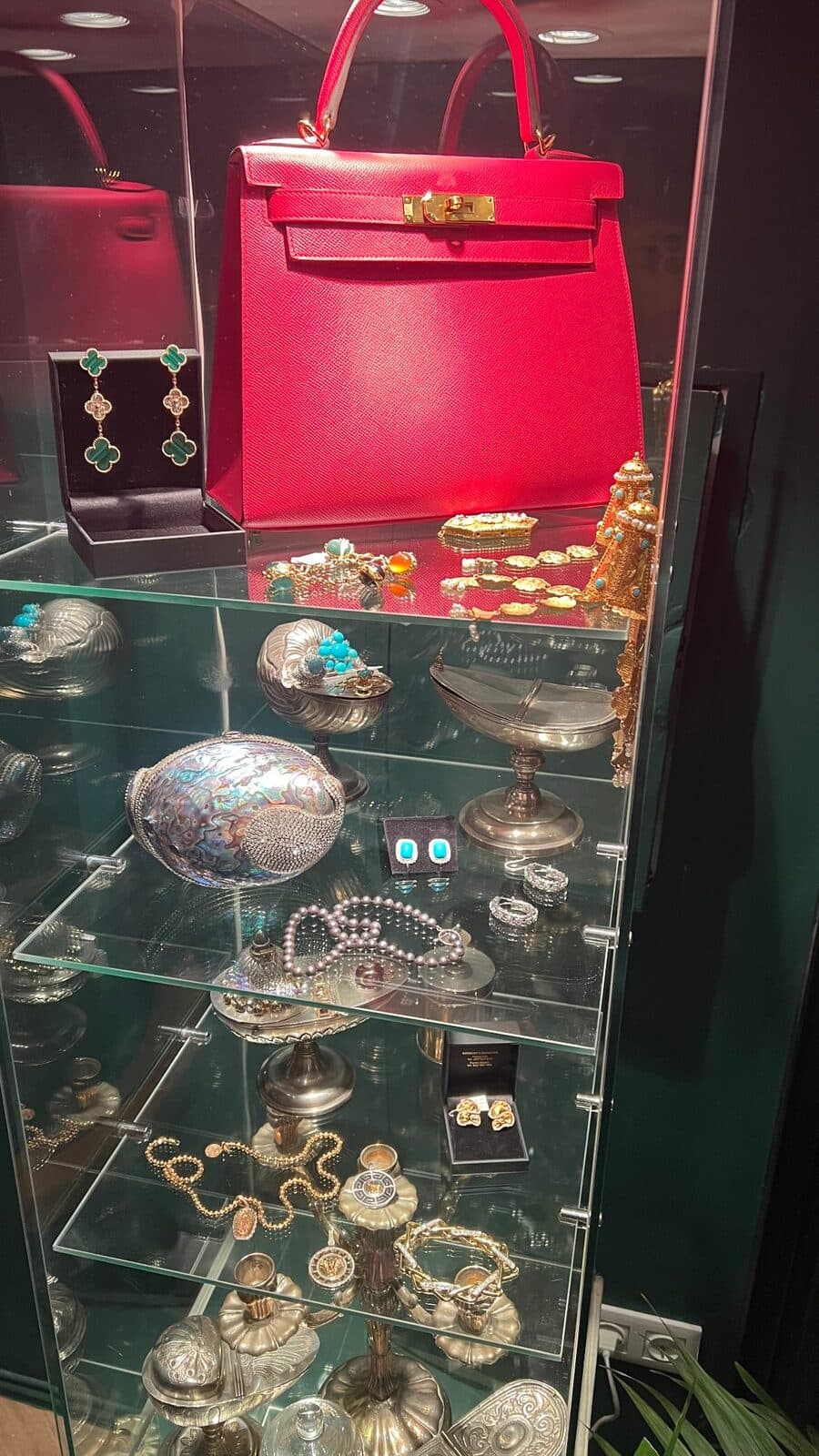 A lit glass cabinet with a red handbag, silverware and jewellery on its shelves.