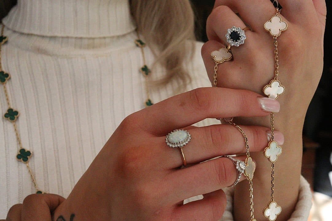 Hands wearing several rings and holding long necklaces against a cream jumper.